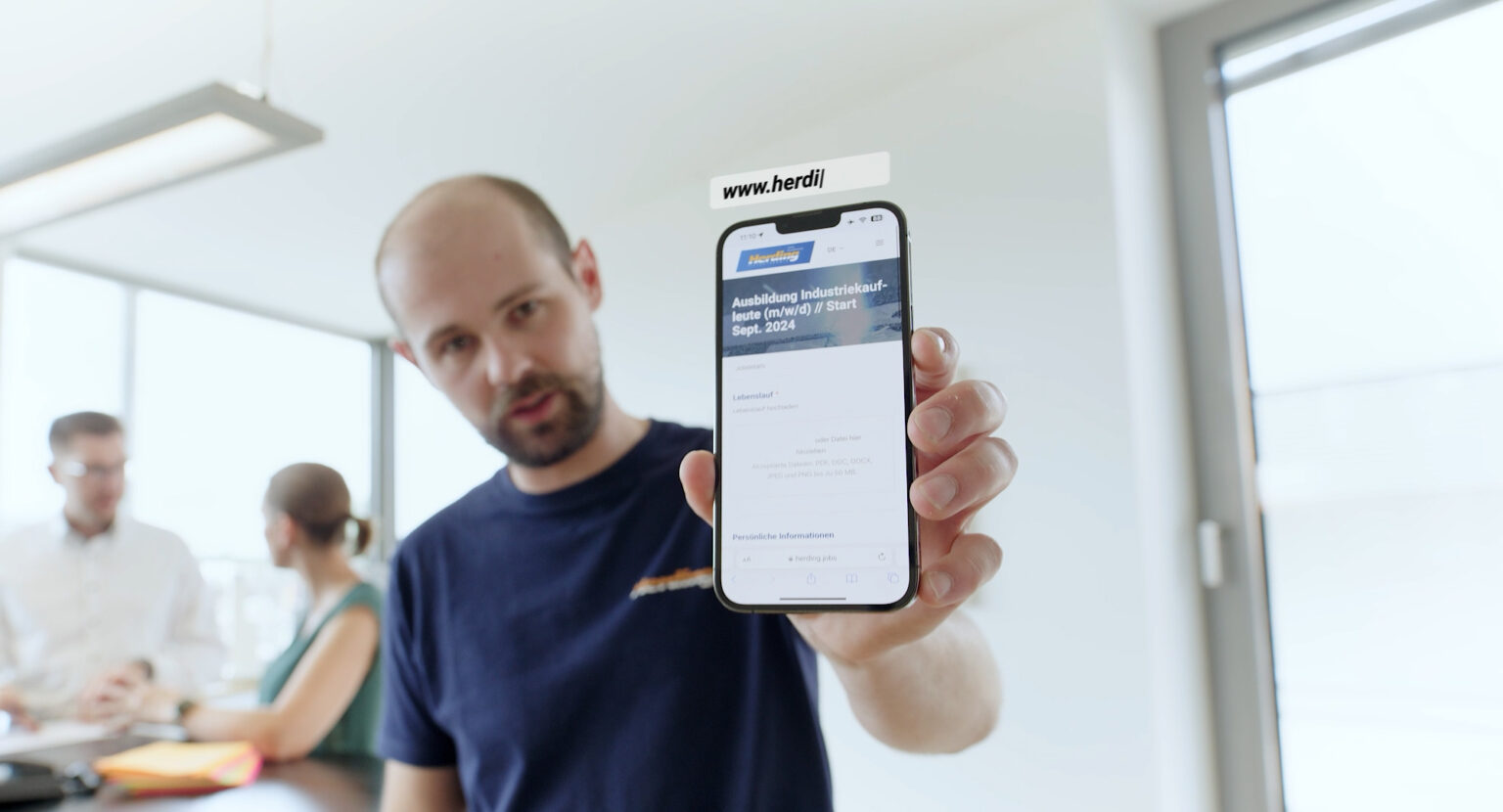 Standbild aus einer Werbung, die einen Mann zeigt, der in einer hell erleuchteten Büroumgebung ein Smartphone in die Kamera hält.